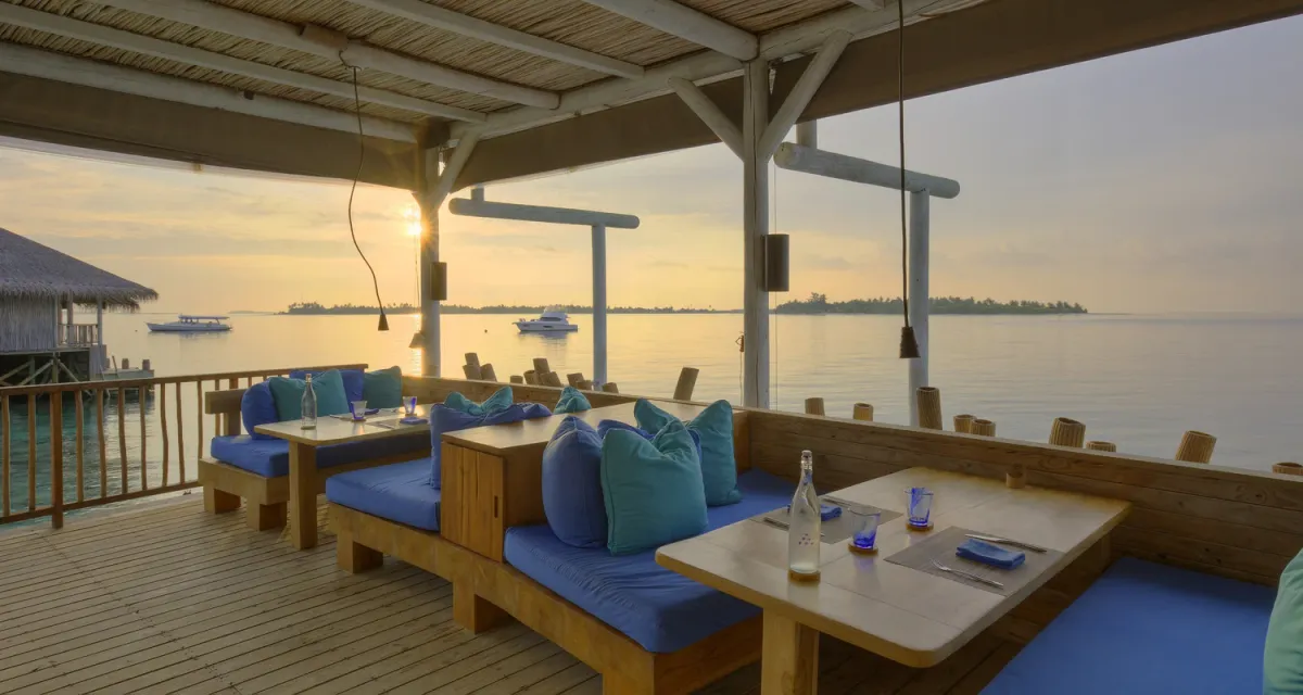 Chill Bar over-water deck with blue cushions facing west at sunset, dive boat moored in the distance at Six Senses Laamu.