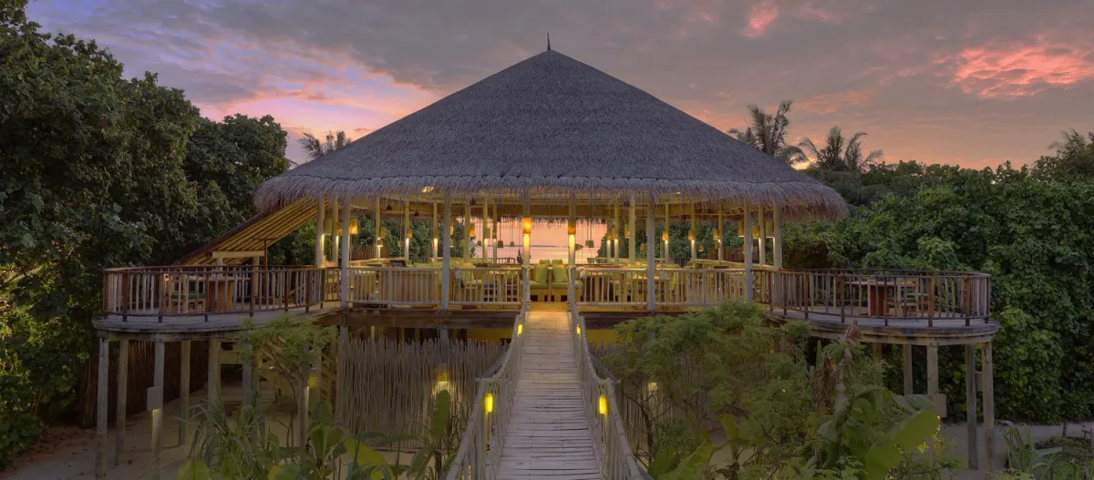 Leaf restaurant, thatched-roof open pavilion on stilts at sunset with boardwalk approach at Six Senses Laamu.