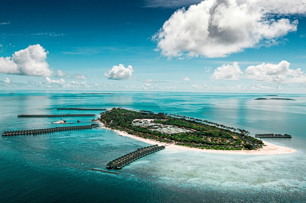 Siyam World Maldives, aerial of Dhigurah island with long overwater villa row extending into the lagoon and jetty with moored boats, Noonu Atoll, Maldives