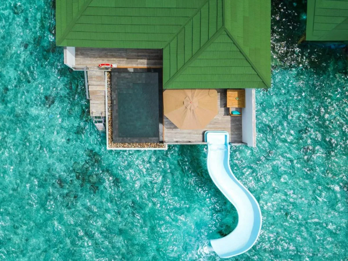 Siyam World Maldives, Water Villa with Pool and Slide top-down aerial showing green-tile roof, plunge pool, parasol deck and curved waterslide into lagoon, Noonu Atoll, Maldives