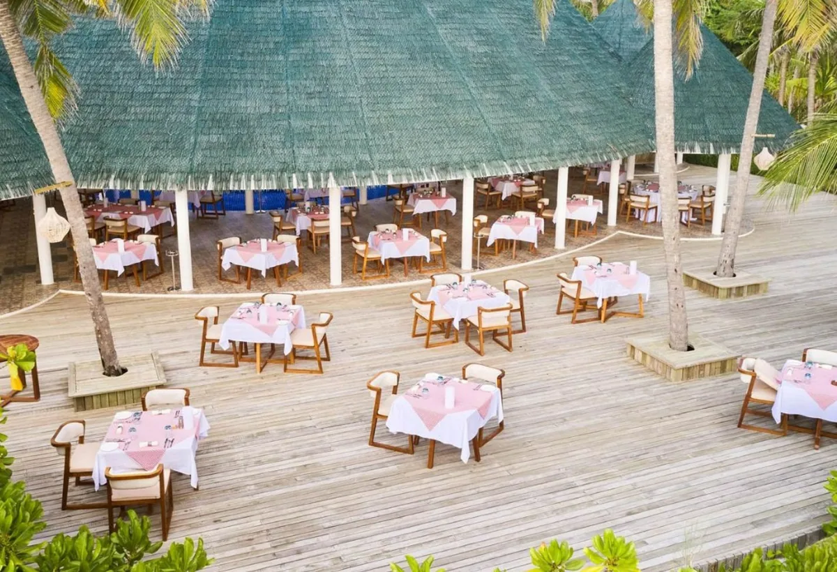 Siyam World Maldives, open-air thatched dining pavilion with pink linen tables, wooden armchairs and coconut palms on beachfront wooden deck, Noonu Atoll, Maldives