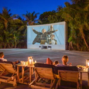 Cinema Paradiso open-air cinema screen rigged among trees with diners seated on the sand at Soneva Fushi.