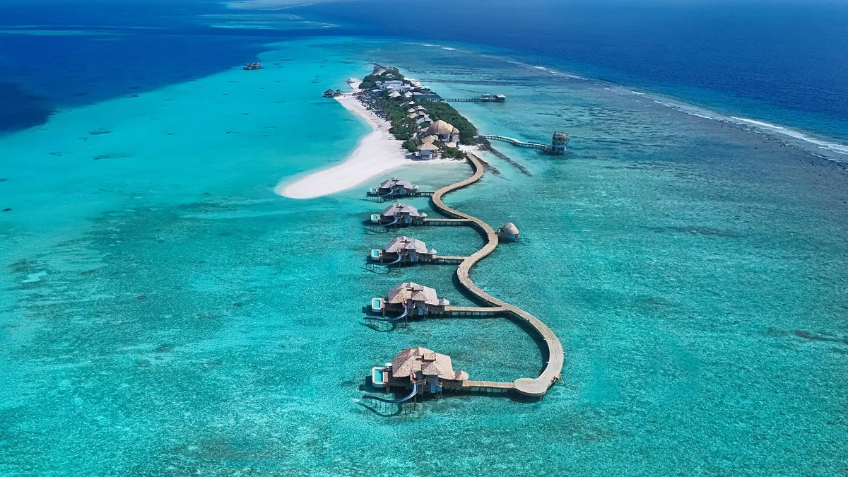 Soneva Secret, aerial, Haa Dhaalu Atoll, Maldives, Soneva Secret drone aerial of Dhipparufushi: the curved overwater jetty trailing from the beach-end main island, overwater villa row along the lagoon's deep-blue drop-off, the Out of This World tower visible on the main island, southwestern Haa Dhaalu Atoll.