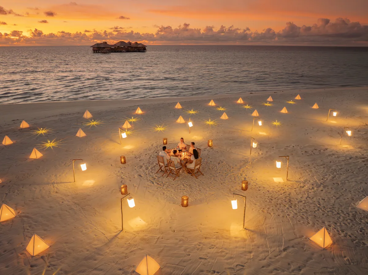 Soneva Secret, dining, Haa Dhaalu Atoll, Maldives, Destination beach dining at sunset: a candlelit private table set on the sand, the property's pyramidal lantern installation lit overhead, the overwater villa row silhouetted across the lagoon in the golden-hour glow.