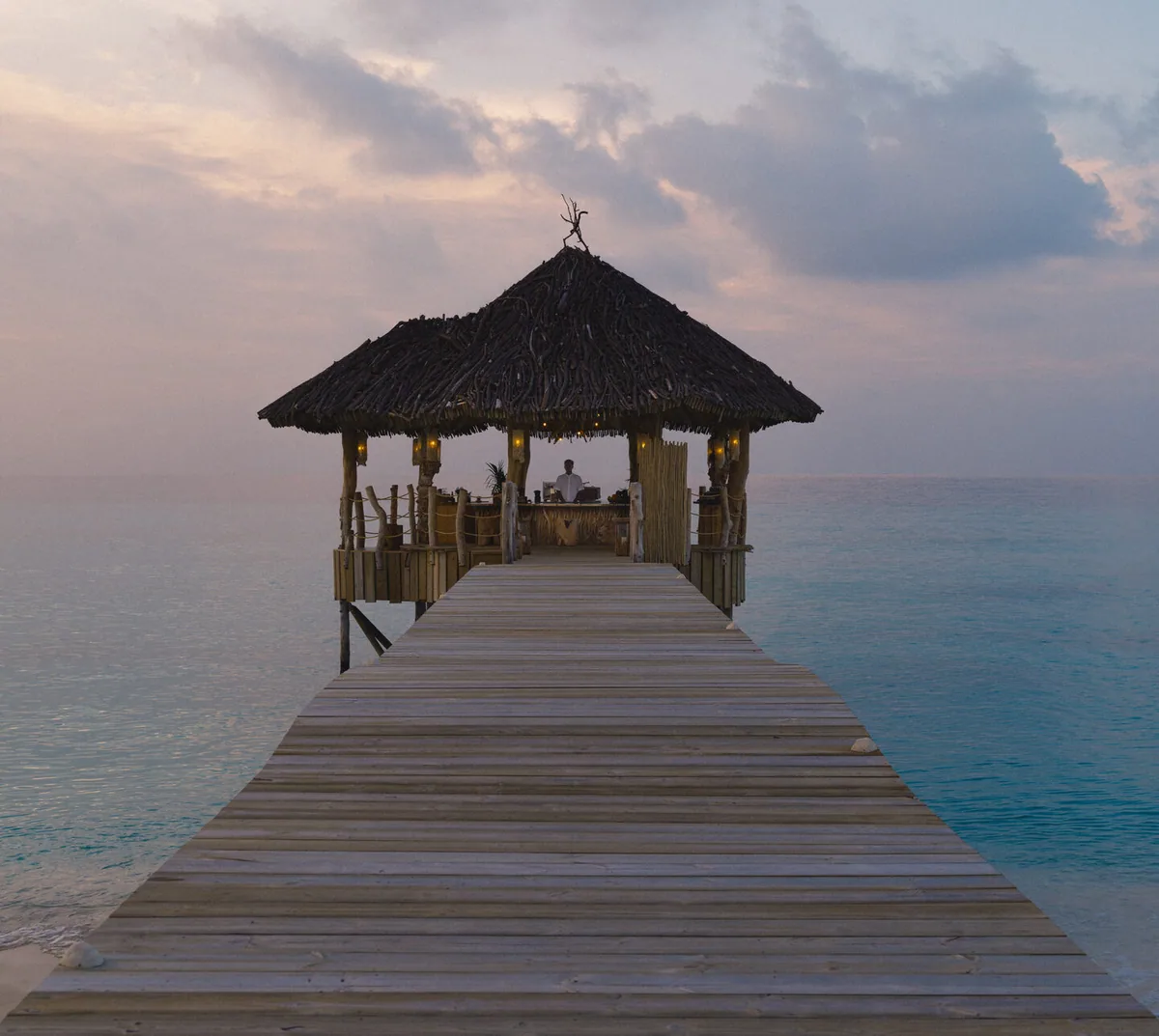 Soneva Secret, dining, Haa Dhaalu Atoll, Maldives, The Secret Bar pavilion at dusk: thatched-roof timber structure at the lagoon's edge, the wooden jetty trailing across the shallow turquoise water to the bar's open deck.