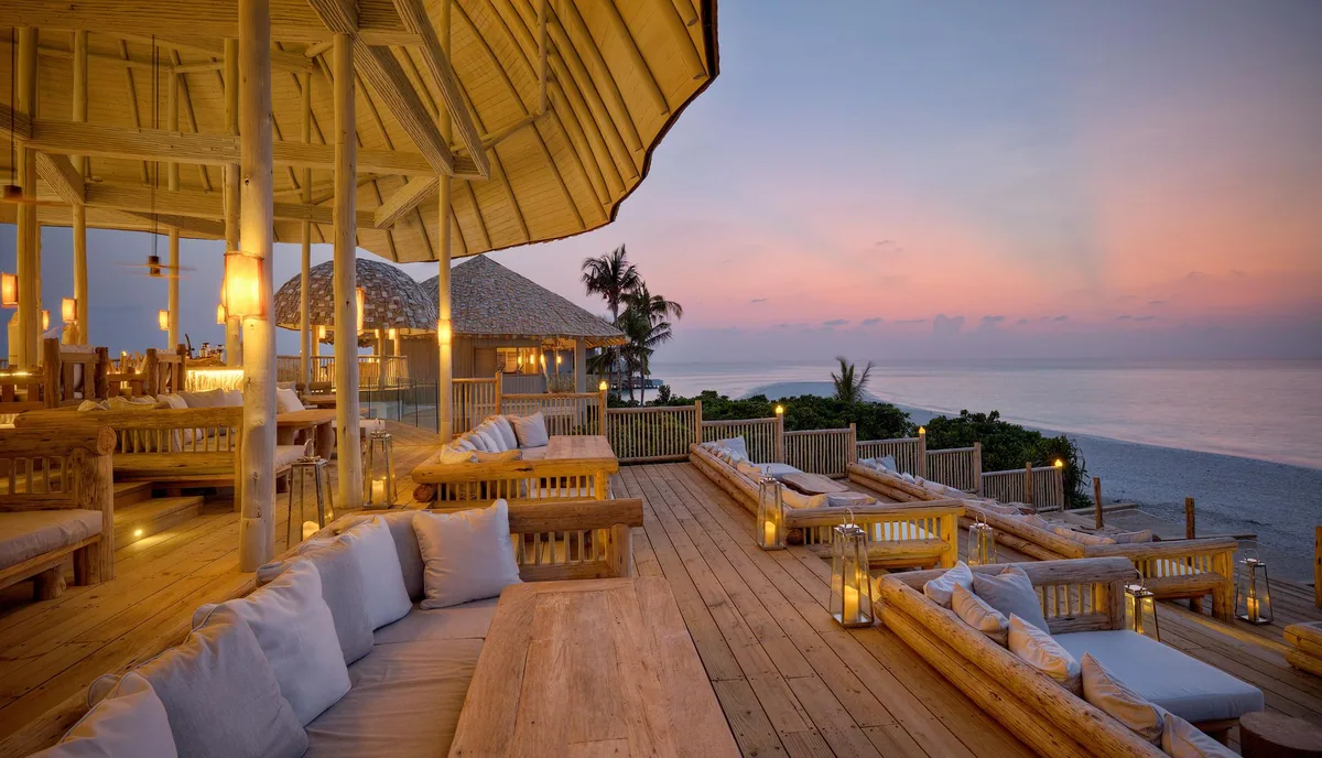 Soneva Secret, dining, Haa Dhaalu Atoll, Maldives, The Den lounge interior at sunset: bamboo-clad walls and ceiling, low daybeds with woven cushions, hanging woven-rattan pendant lanterns casting warm light across the open-air cocktail and music venue.