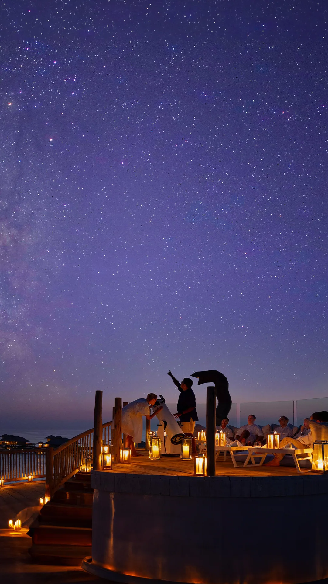 Soneva Secret, activities, Haa Dhaalu Atoll, Maldives, The observatory deck under the Milky Way: a guide pointing out a constellation beside the telescope, guests seated at candlelit tables on the timber deck, the night-sky programme that anchors the Out of This World astronomy configuration.