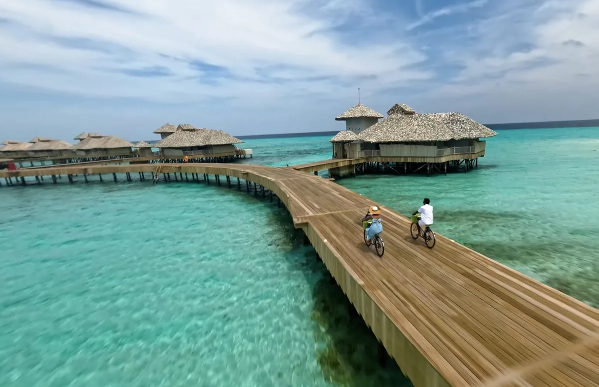 Soneva Secret, activities, Haa Dhaalu Atoll, Maldives, A couple cycling along the timber over-water jetty connecting the thatched-roof villas, the turquoise lagoon directly underneath the boardwalk, daylight Soneva-island life detail.