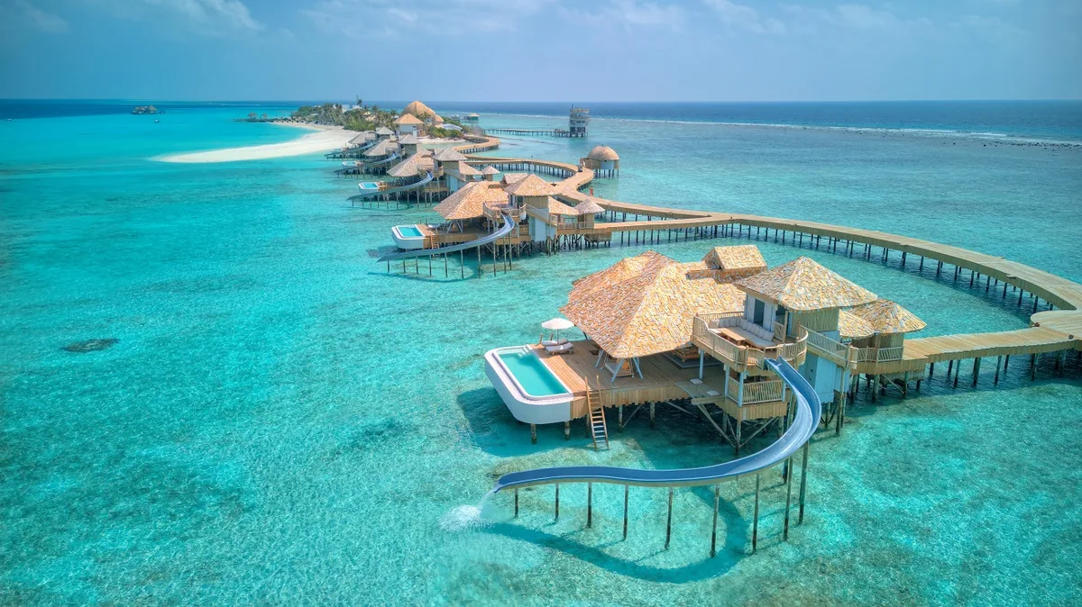 Soneva Secret, villa overwater, Haa Dhaalu Atoll, Maldives, Top-down aerial of the over-water villa cluster: the curving jetty rising from the sandbank with the thatched-cone-roofed Horizon villas stepping out across the turquoise lagoon shallows.
