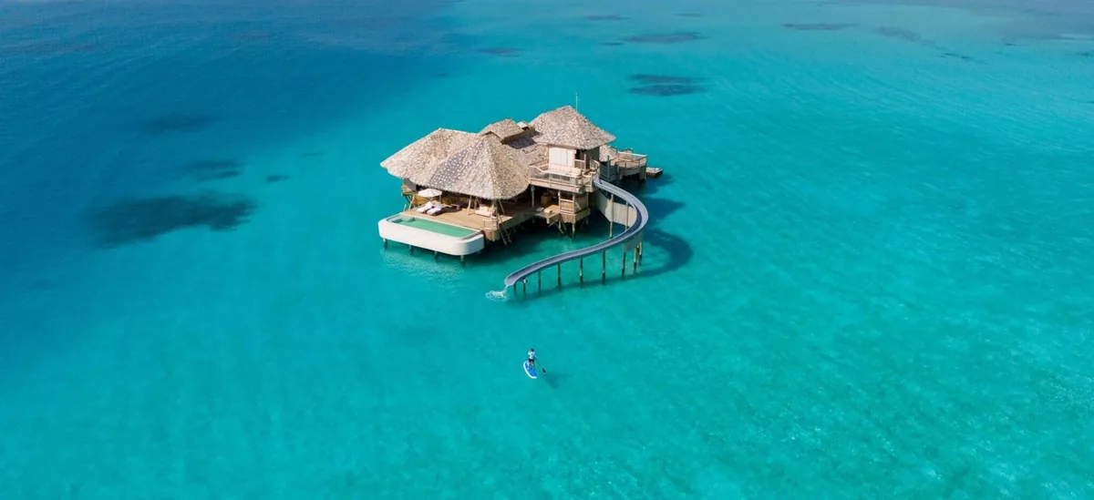 Soneva Secret, villa overwater, Haa Dhaalu Atoll, Maldives, Aerial of the Overwater Hideaway villa: thatched cone roofs, the signature lagoon-entry water slide curling from the villa deck, a paddleboarder gliding across the reef-shallow lagoon below.
