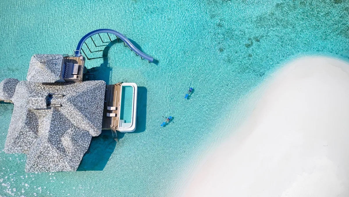 Soneva Secret, activities, Haa Dhaalu Atoll, Maldives, Top-down aerial of an Overwater Hideaway villa: thatched-cone roof, lagoon-entry slide, and two glass-bottomed kayaks paddling alongside the sand-fringed villa edge.