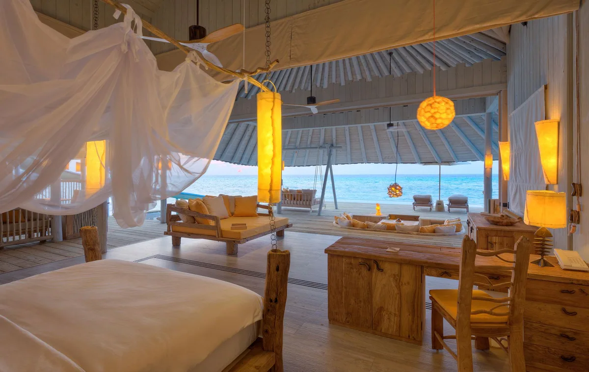 Soneva Secret, villa overwater, Haa Dhaalu Atoll, Maldives, Horizon Overwater Villa interior at dusk: four-poster bed with mosquito net, driftwood pendant lanterns, organic-timber furniture, the retractable wall slid back to reveal the deck with hanging daybed swings and the lagoon beyond.