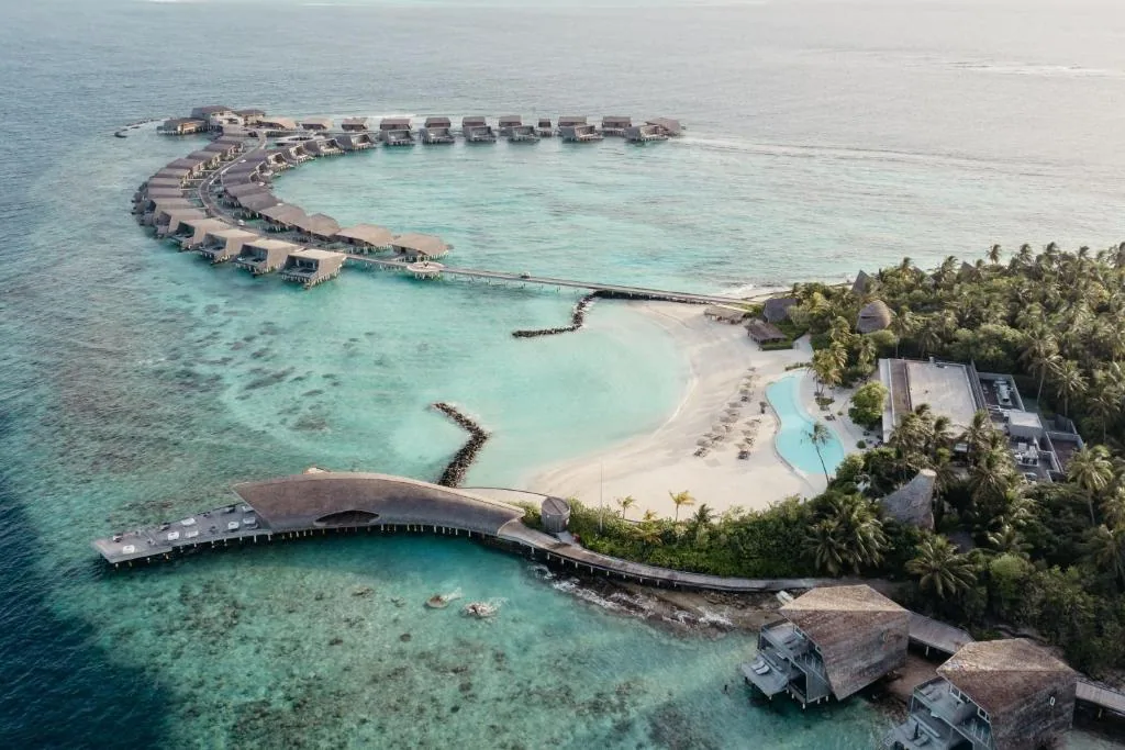 The St. Regis Maldives Vommuli Resort, aerial, Dhaalu Atoll, Maldives, Property overview aerial: the curved Whale Bar arrival walkway sweeps off the main island, framing the central beach, the swimming pool, and the C-shaped overwater villa ring that wraps the southern reef edge. The geometry that gives Vommuli its house style in a single frame.