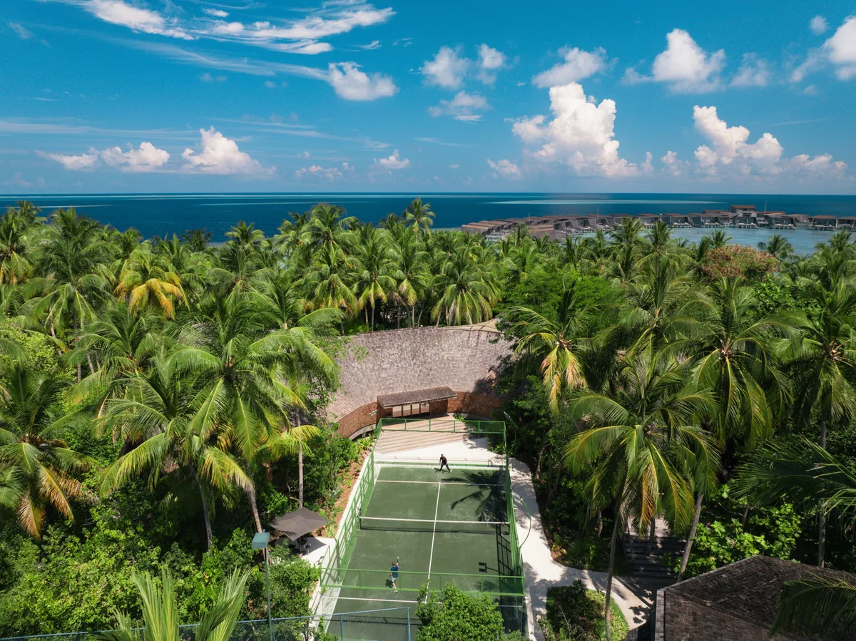 The St. Regis Maldives Vommuli Resort, activities, Dhaalu Atoll, Maldives, Padel court aerial: the resort's lighted hard court tucked into the palm-canopy interior of the island, a thatched pavilion shading the spectator side, the overwater villa cluster reading as a brown ribbon along the lagoon edge in the distance. The recent activity-programme addition shown at scale.