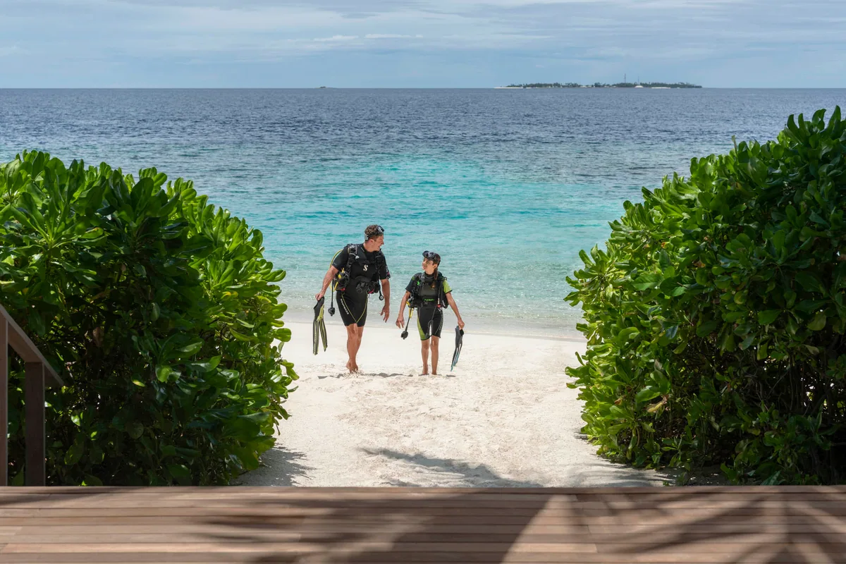 The St. Regis Maldives Vommuli Resort, activities, Dhaalu Atoll, Maldives, Dive exit on the property beach: two divers in wetsuits walk through a gap in the hedge line onto the white-sand crescent, fins in hand, the deep-water adjacent island visible in the far distance. The PADI operation's standard entry point on the southern reef side.