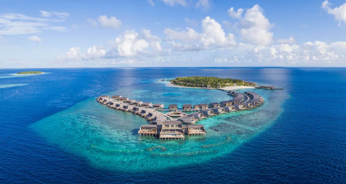 The St. Regis Maldives Vommuli Resort, aerial, Dhaalu Atoll, Maldives, Wider aerial: the overwater village in the foreground reads as two arms of villas meeting at a central pavilion, the main palm-canopied island sitting beyond, the reef shelf visible as a paler turquoise ring before the open ocean. Reef geometry and villa-cluster scale in one view.