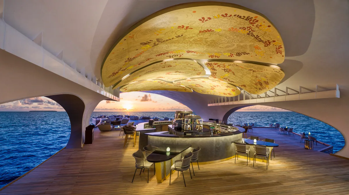 The St. Regis Maldives Vommuli Resort, dining, Dhaalu Atoll, Maldives, Whale Bar inside the Vommuli House clubhouse: the painted whale-belly underside, all warm gold and coral motifs, arches over a circular polished-steel central bar, the timber deck running through to a sunset-facing ocean edge. The country's only major non-rectangular over-water venue.