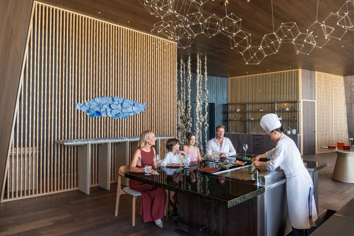 The St. Regis Maldives Vommuli Resort, dining, Dhaalu Atoll, Maldives, Orientale Cantonese chef's counter: a constellation of geometric brass pendants overhead, slatted timber walls, a wave-like ceramic art panel, the chef plating to a four-top counter that reads as the venue's signature seat. The tea-ceremony-pairing room at its tightest configuration.