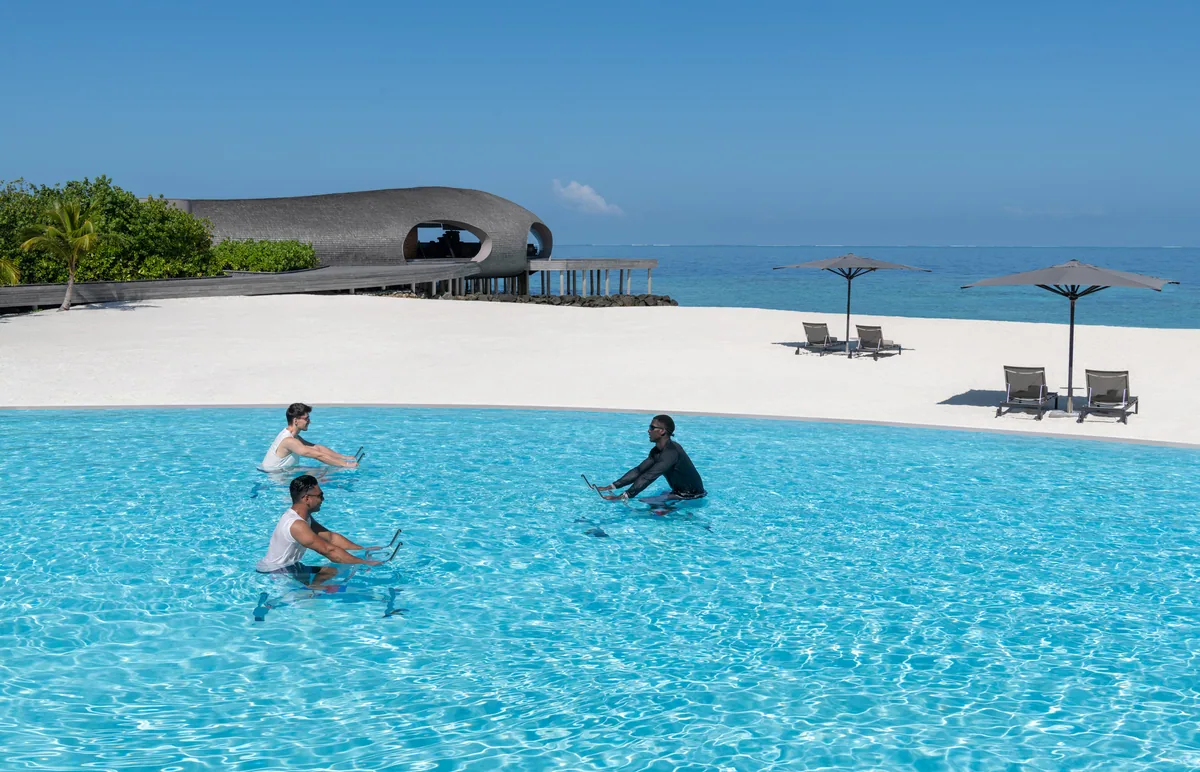 The St. Regis Maldives Vommuli Resort, activities, Dhaalu Atoll, Maldives, Main pool aqua-bike class: three guests on submerged bikes work in the infinity pool while the iconic ribbed-concrete Vommuli House clubhouse sits on the lagoon edge behind, a single jetty walkway running off to the right. Property identity anchored in the background frame.