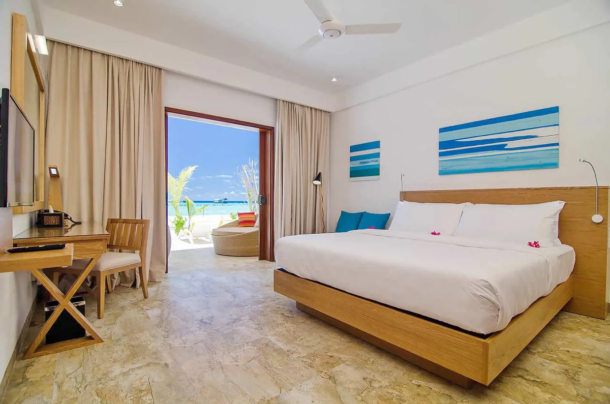 Summer Island Maldives, villa beach, North Malé Atoll, Maldives, Beach Villa bedroom interior: king bed dressed in white linens with turquoise pillows and pink-flower garnish, light-wood platform bed frame, abstract striped blue-and-white wall art panels above the bed, beige curtains drawn back, open glass door framing palms and the lagoon jetty beyond, warm marble-pattern tile floor.