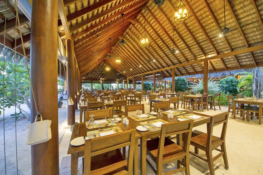 Summer Island Maldives, dining, North Malé Atoll, Maldives, Samuga restaurant: open-sided pavilion with thatched pitched roof and exposed wood-beam ceiling, dark-iron pendant lantern cluster, dark-wood tables and chairs set with napkins and crystal glassware, sand floor at the perimeter, tropical foliage framing the open edges.