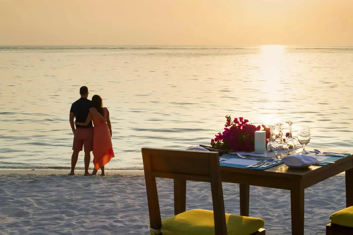 Summer Island Maldives, dining, North Malé Atoll, Maldives, Sunset private beach dinner staging: couple at the shoreline silhouetted against the sun on the horizon, dinner table set on the sand with magenta-bougainvillea centrepiece, candles and crystal glassware, yellow-cushioned timber chairs, glassy lagoon reflecting the warm dusk light.
