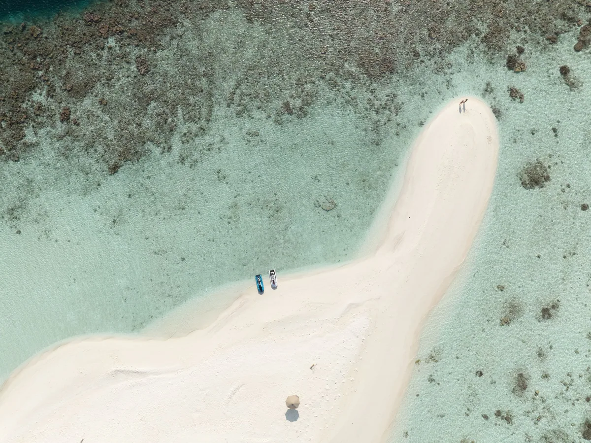 Sun Siyam Olhuveli, aerial, South Malé Atoll, Maldives, Top-down aerial of a tear-drop-shaped sandbank within the property's lagoon network: two figures standing at the sandbank tip, twin kayaks beached on the spit, a single sand parasol, the surrounding reef bed shading the lagoon emerald to dark turquoise.
