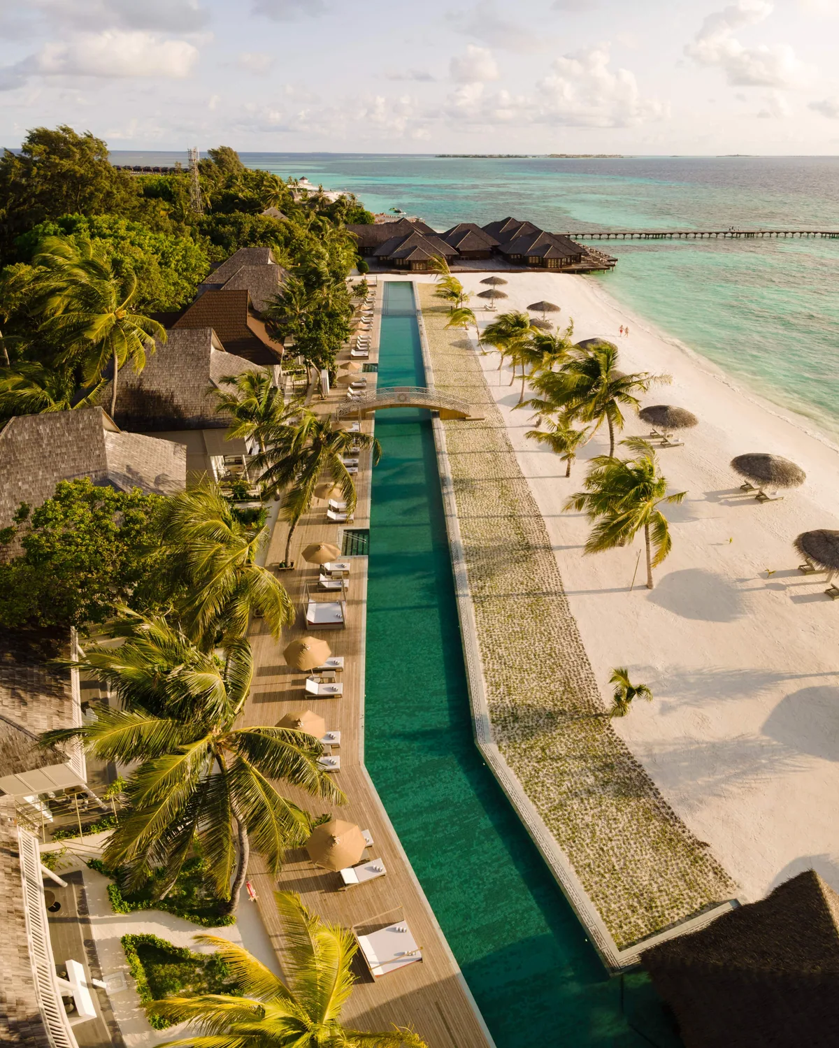 Sun Siyam Olhuveli, aerial, South Malé Atoll, Maldives, Angled aerial pano of the long infinity pool and beach: tall coconut palms framing the foreground, ranks of sun-loungers along the pool edge, the over-water villa boardwalk extending into the lagoon at the top of frame, the resort's mid-range scale visible in a single composition.