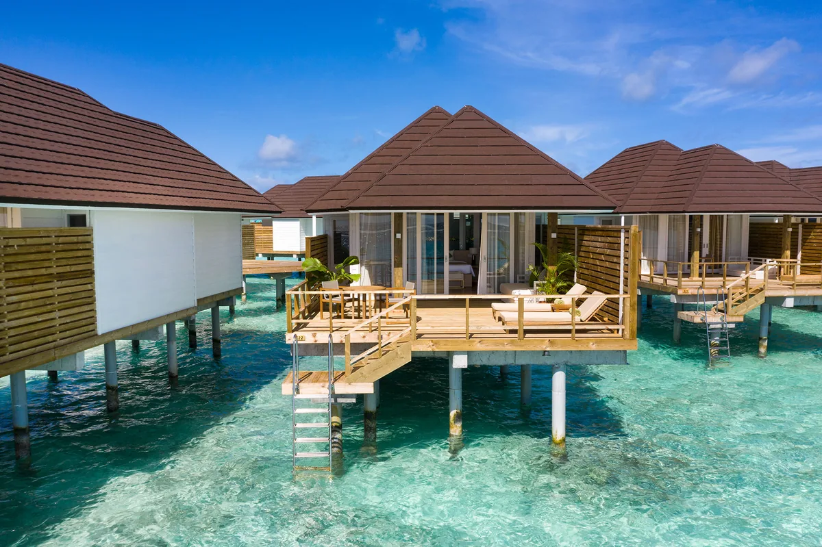 Sun Siyam Olhuveli, villa overwater, South Malé Atoll, Maldives, Grand Water Villa exterior viewed from the lagoon: brown plank construction with dark shingle peaked roof, white interior glimpsed through sliding doors, wood-screen privacy wing, twin loungers on the deck, a lagoon ladder dropping into the crystal water below.