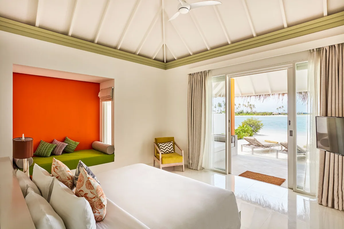 Sun Siyam Olhuveli, villa beach, South Malé Atoll, Maldives, Grand Beach Villa interior at the contemporary-bright tier: peaked white ceiling, a vibrant orange-and-green window-seat banquette anchoring the room, yellow chevron-print armchair, white tile floor, sliding glass doors opening to a private deck with twin loungers and the palm-fringed lagoon beyond.