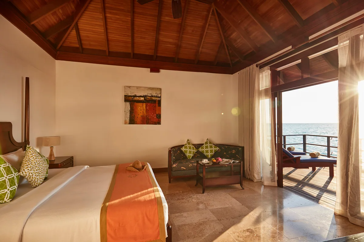 Sun Siyam Olhuveli, villa overwater, South Malé Atoll, Maldives, Water Villa interior at the traditional-warm tier: dark exposed-beam peaked ceiling, beige stone tile floor, neutral palette with green-and-orange accent throws, dark-wood furniture, French doors framing afternoon sun streaming onto the deck and lagoon beyond.