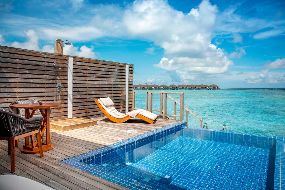 Sun Siyam Olhuveli, villa honeymoon, South Malé Atoll, Maldives, Honeymoon Romantic Water Villa with private plunge pool: blue-tile pool set into the deck with infinity edge over the lagoon, twin orange-cushion deck loungers, wood-screen privacy wall, outdoor shower stall, a dining table for two, the adjacent over-water villa row visible across the water at the Honeymooners-section wing.