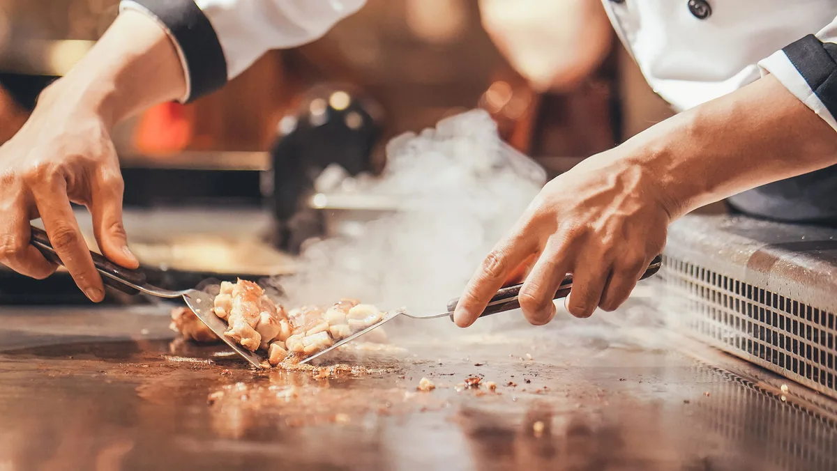 Sun Siyam Olhuveli, dining, South Malé Atoll, Maldives, Four Spices teppanyaki action close-up: chef hands working twin spatulas across the flat grill, a portion of meat seared with steam rising into the frame, the documentary face of the Japanese-Indian-Thai-Maldivian specialty venue.