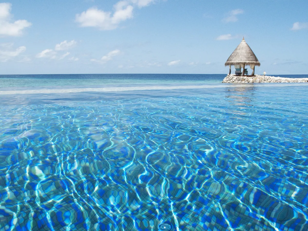 Taj Coral Reef Resort & Spa, setting, North Malé Atoll, Maldives, Lagoon-edge infinity pool with the crystal-blue water plane merging into the open lagoon at the visual horizon, a small thatched gazebo at the breakwater rocks on the right, and a wisp of cloud over the open ocean beyond.