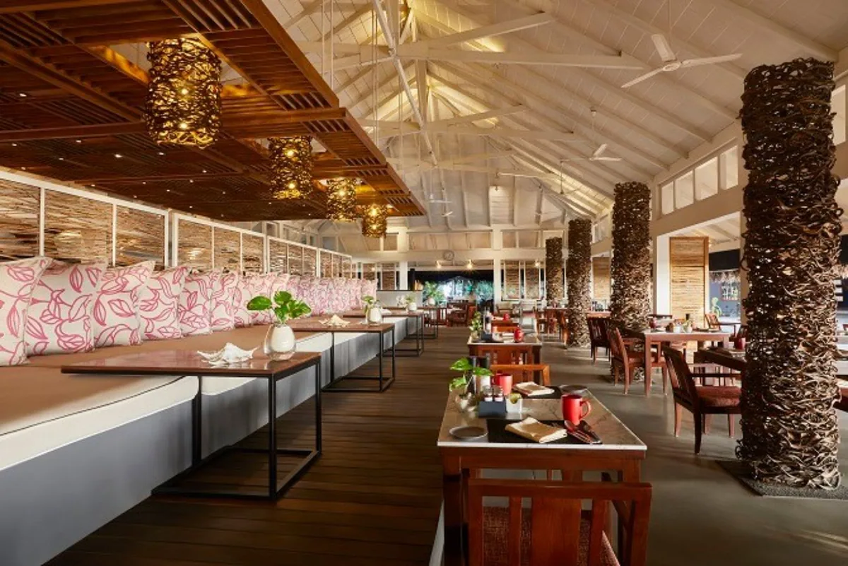 Taj Coral Reef Resort & Spa, dining, North Malé Atoll, Maldives, Bokkura main restaurant interior, the high pitched white ceiling with timber rafter structure, rope-wrapped column treatments, woven wicker pendant lights, pink floral throw cushions along the long banquette, and wood-plank flooring (the trendy modern setting for the Continental + Indian + Pan-Asian + Sri Lankan + Maldivian fusion menu).