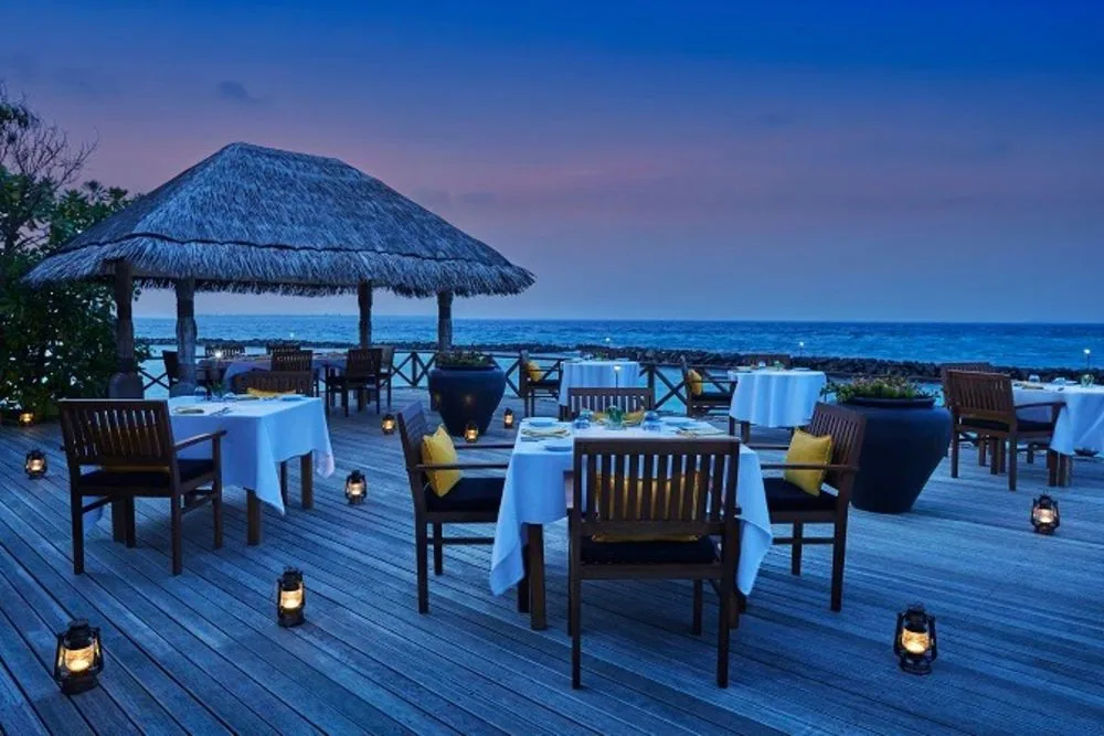 Taj Coral Reef Resort & Spa, dining, North Malé Atoll, Maldives, Open The Grill outdoor deck venue at twilight, the thatched-roof pavilion against the pink-blue dusk sky, white-linen tables with yellow accent cushions, hurricane lanterns lining the timber deck, and the breakwater-edged lagoon as backdrop.