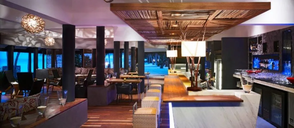 Taj Coral Reef Resort & Spa, dining, North Malé Atoll, Maldives, Reef Bar interior, the white-marble counter with backlit cobalt-blue bar display, the central bamboo-tree decorative installation with pendant box-shades suspended above, the woven rattan ceiling lattice panel, and lounge seating with floral throw cushions opening to the lagoon.