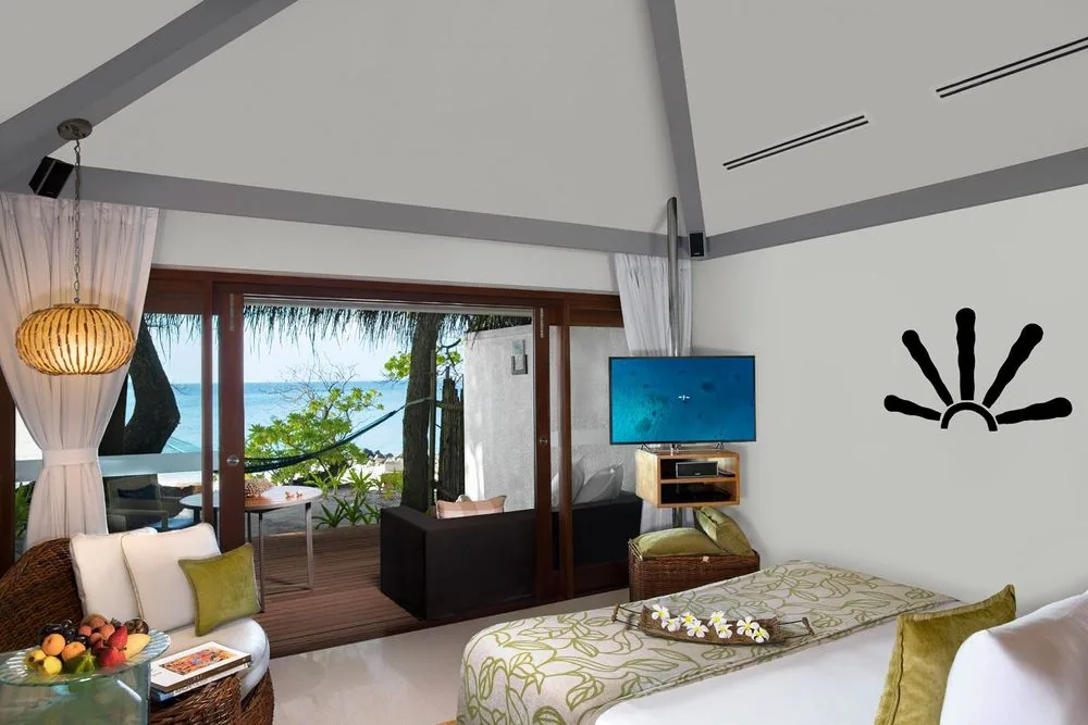 Taj Coral Reef Resort & Spa, villa beach, North Malé Atoll, Maldives, Superior Charm Villa interior, the pitched white ceiling with grey timber rafter accents, the signature starburst medallion in black on the accent wall (matching the over-water villa motif), woven rattan pendant light, retractable timber-framed sliding doors opening to a hammock-strung veranda and beach view, and an olive-green floral runner with frangipani arrangement on the bed.
