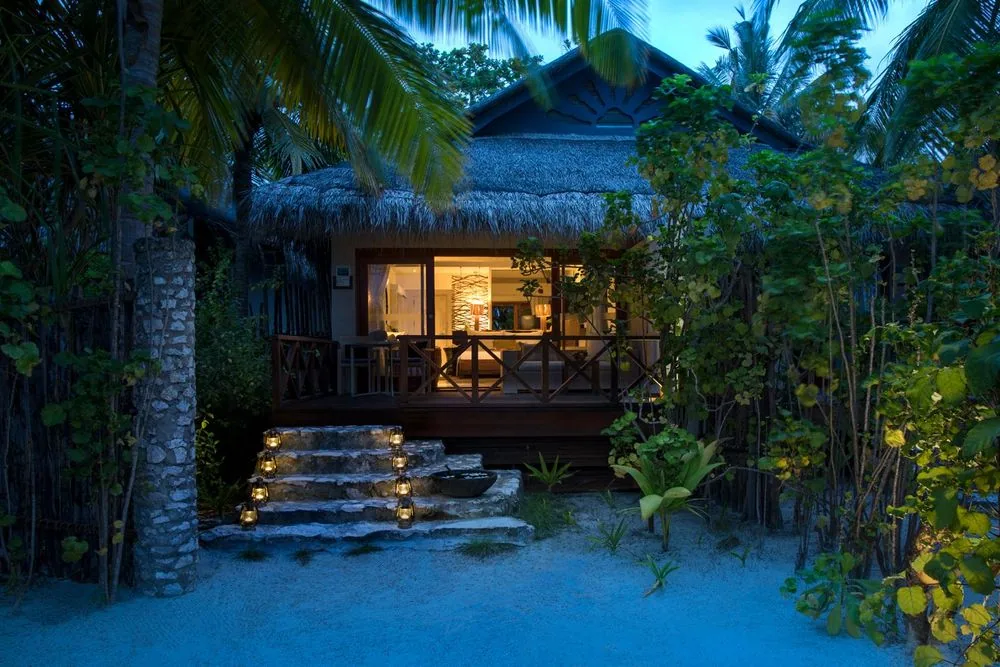 Taj Coral Reef Resort & Spa, villa beach, North Malé Atoll, Maldives, Deluxe Charm Villa exterior at twilight, the standalone thatched-pitched-roof bungalow nestled into the jungle vegetation, the warm-lit interior glow through the timber-framed glazing, a covered veranda with stepped coral-stone approach, and hurricane lanterns lining the path through the palm canopy.