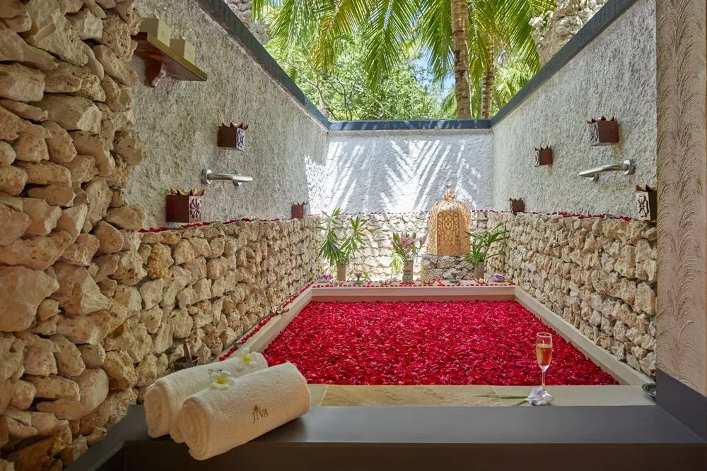 Taj Coral Reef Resort & Spa, spa, North Malé Atoll, Maldives, Jiva Spa outdoor treatment garden, the coral-stone perimeter walls, the rose-petal-filled bath as treatment centrepiece, two brass wall-mounted water spouts under decorative lattice panels, a slim ledge with frangipani arrangements at the far wall, and rolled white towels with the embroidered JIVA wordmark in the foreground.
