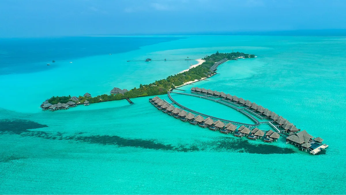 Taj Exotica Resort & Spa, Maldives, aerial, South Malé Atoll, Maldives, Wide aerial of the full Emboodhu Finolhu island geometry in northern South Malé Atoll, with the curved overwater villa cluster extending to the right, a smaller cluster at left, and the palm-canopy main island spine carrying the property's central infrastructure.