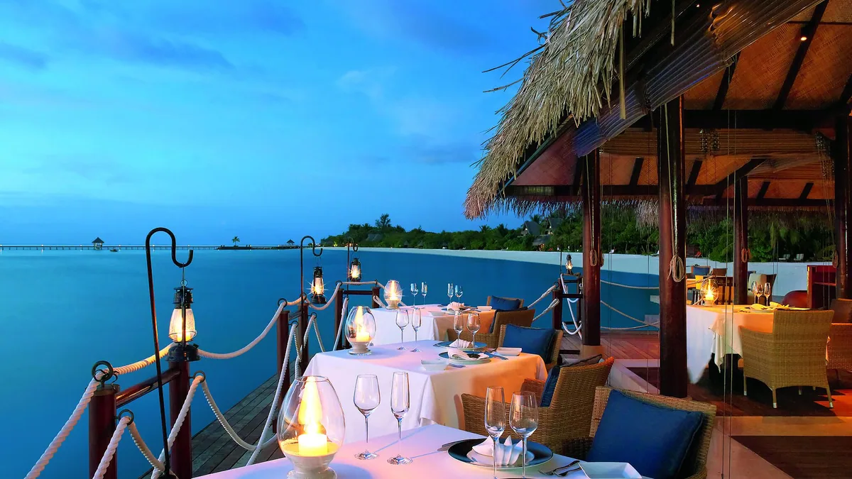 Taj Exotica Resort & Spa, Maldives, dining, South Malé Atoll, Maldives, Deep End over-water specialty venue at blue hour, candle-lit white tablecloths and crystal stemware on the open deck, brass hurricane lanterns hung from rope-and-stanchion balustrades, thatched canopy at right, calm twilight lagoon and distant property jetty on the horizon.