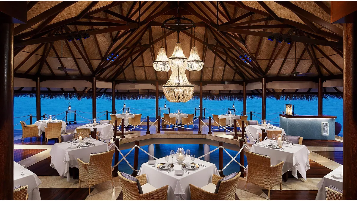 Taj Exotica Resort & Spa, Maldives, dining, South Malé Atoll, Maldives, Deep End restaurant interior, vaulted timber pavilion with a single large crystal chandelier suspended above concentric tables that ring a central open well looking down into the lagoon (the venue's signature feature), thatched-eave openings to the open lagoon, rope-balustrade dividers between table tiers.