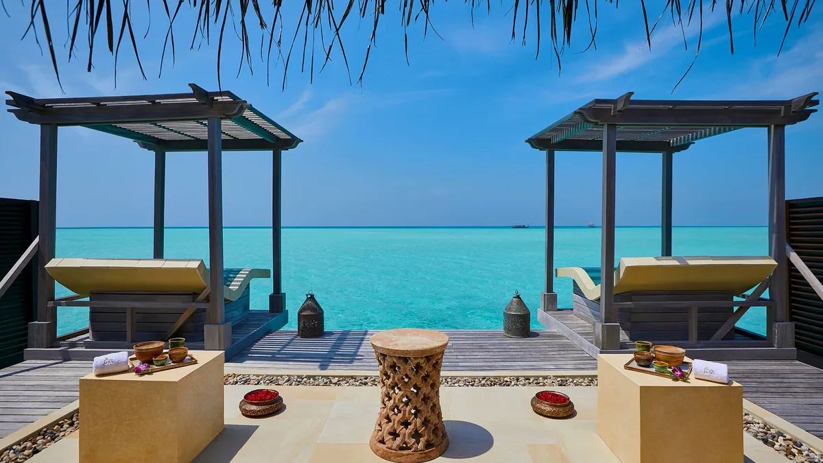 Taj Exotica Resort & Spa, Maldives, spa, South Malé Atoll, Maldives, Jiva Spa open-air relaxation deck, twin timber pergolas over the lagoon with cushioned daybeds, traditional Indian carved wooden stool and copper bowls (Ayurvedic ritual props) on the threshold platform, brass lanterns at the perimeter.