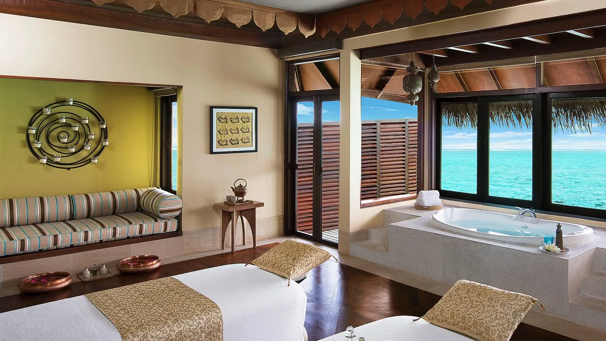 Taj Exotica Resort & Spa, Maldives, spa, South Malé Atoll, Maldives, Jiva Spa overwater couple treatment room interior, twin massage tables in the foreground, a soaking jacuzzi set against the ocean-view window, carved-timber wall art and pierced-metal Indian lanterns at the corners, daybed nook with striped cushions in the recess.
