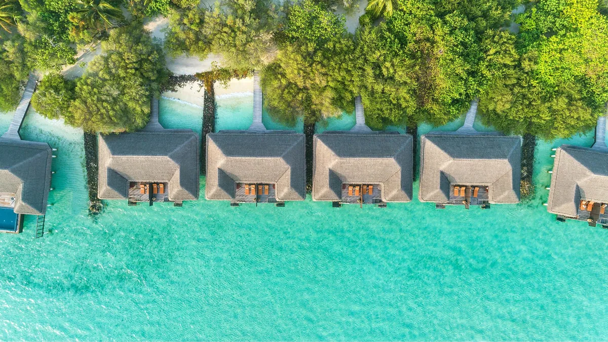 Taj Exotica Resort & Spa, Maldives, aerial, South Malé Atoll, Maldives, Top-down aerial of a row of six thatched-roof beach-villa pavilions along the lagoon perimeter, palm-canopy backdrop and turquoise reef-flat foreground at the perimeter beach configuration.