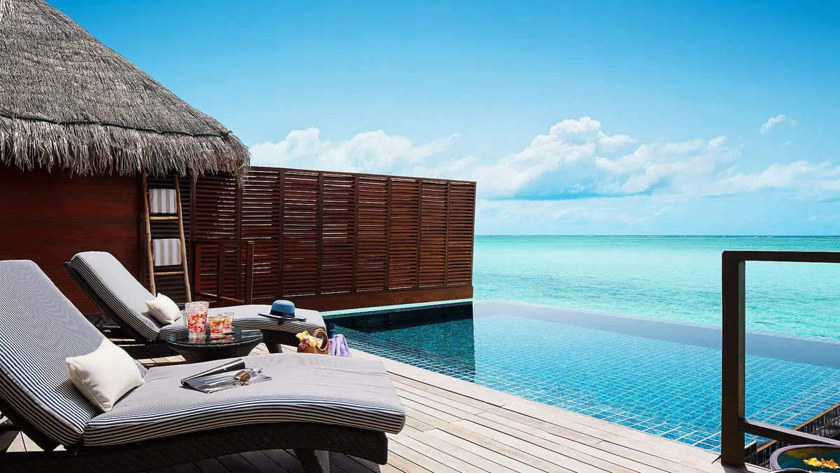Taj Exotica Resort & Spa, Maldives, villa overwater, South Malé Atoll, Maldives, Ocean Suite deck close-up, striped sun loungers, dark louvered villa screen, infinity-edge plunge pool spilling into the ocean horizon at the over-water configuration.