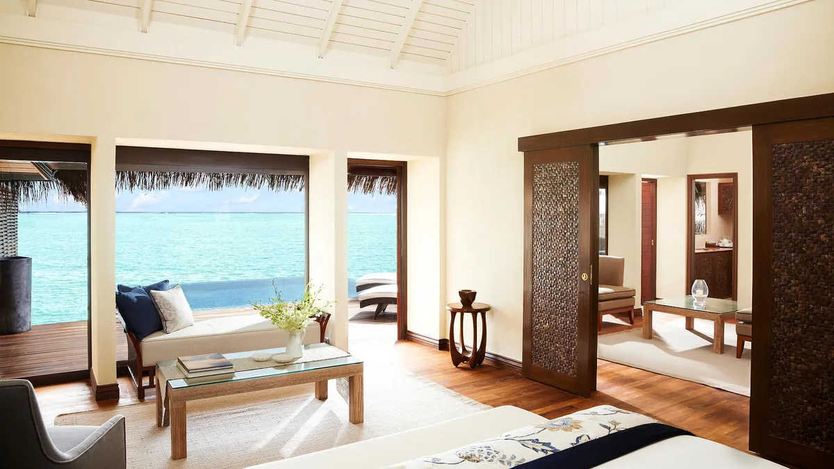 Taj Exotica Resort & Spa, Maldives, villa interior, South Malé Atoll, Maldives, Overwater villa interior with vaulted white-timber ceiling, hardwood floors, daybed beside a picture-frame window framing turquoise lagoon, carved wooden screen door at the en-suite threshold.