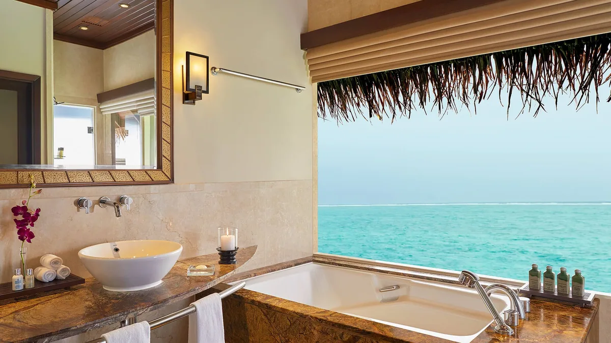 Taj Exotica Resort & Spa, Maldives, villa interior, South Malé Atoll, Maldives, Deluxe Lagoon Villa bathroom, brown-marble double vanity and a soaking tub set against a picture-frame window with thatched palm fringe and open-lagoon view, orchid and bath amenities at the sink.