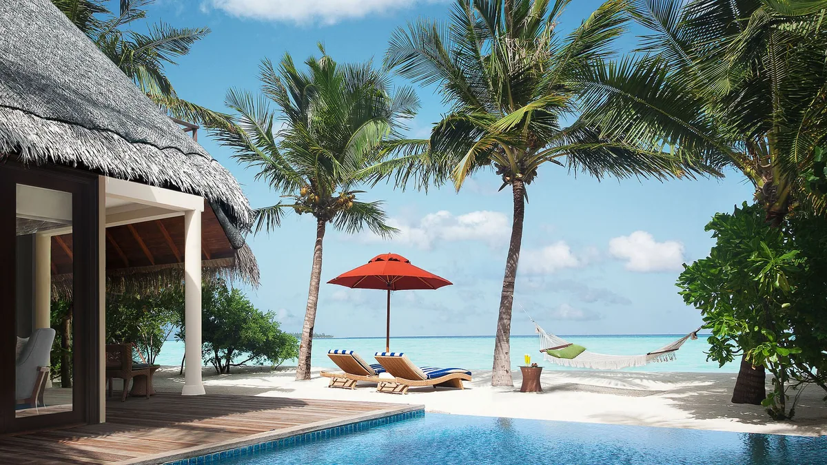 Taj Exotica Resort & Spa, Maldives, villa beach, South Malé Atoll, Maldives, Beach Suite deck with red umbrella shading wood-frame sun loungers, a green hammock strung between palms, private pool in foreground, white-sand frontage opening to the lagoon.